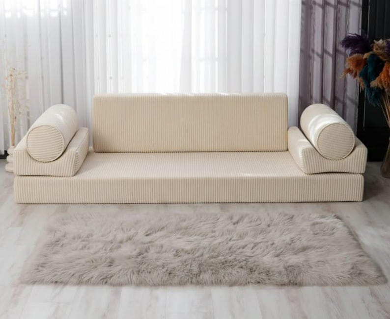 Op de afbeelding: Een beige modulaire bank met een ribfluwelen textuur, bestaande uit een rechthoekige basis, rugleuning en twee cilindrische kussens. Een zacht, grijs rechthoekig vloerkleed ligt voor de bank. De kamer heeft een neutraal kleurenpalet.