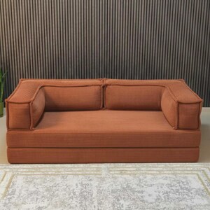 May include: A terracotta corduroy sofa with two back cushions and armrests. The sofa is on a low platform and is placed on a patterned rug. The background features a vertical wood panel wall.
