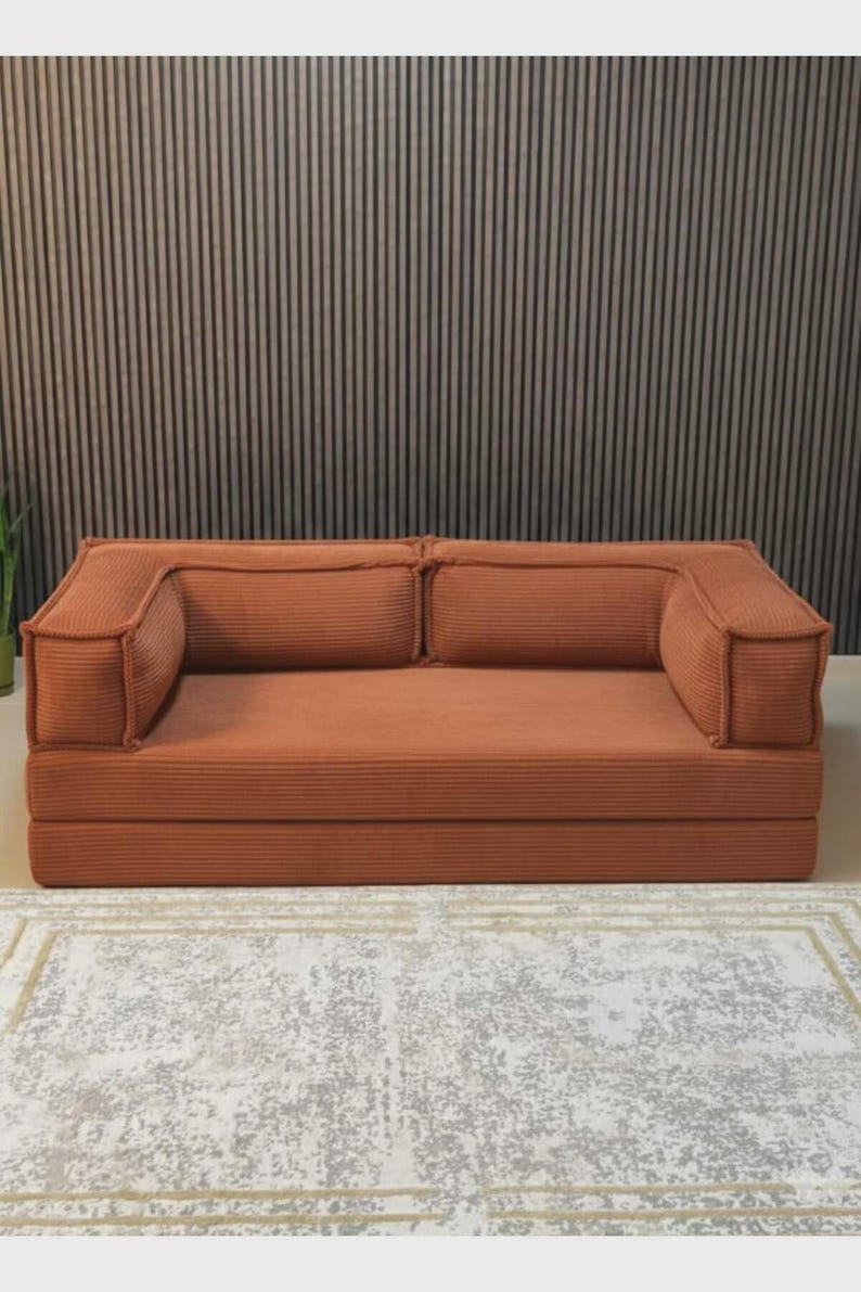 May include: A terracotta corduroy sofa with two back cushions and armrests. The sofa is on a low platform and is placed on a patterned rug. The background features a vertical wood panel wall.