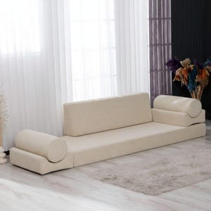 Puede incluir: Sofá de pana beige con un perfil bajo. El sofá tiene una base rectangular, un respaldo y dos reposabrazos cilíndricos. La tela tiene líneas verticales. El sofá está frente a una ventana con cortinas transparentes.