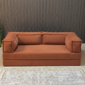 May include: A terracotta corduroy sofa with two back cushions and armrests. The sofa is on a low platform and sits on a patterned rug. The background features a vertical wood panel wall.