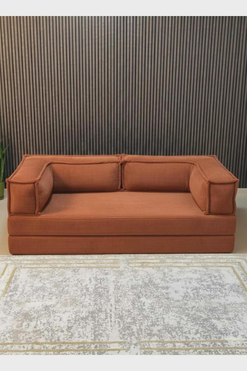 May include: A terracotta corduroy sofa with two back cushions and armrests. The sofa is on a low platform and sits on a patterned rug. The background features a vertical wood panel wall.