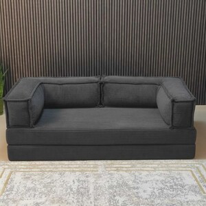 Peut inclure: Un canap&eacute; en velours c&ocirc;tel&eacute; gris fonc&eacute; avec deux accoudoirs et deux coussins de dossier. Le canap&eacute; repose sur une base pli&eacute;e. Le canap&eacute; est sur un tapis &agrave; motifs.