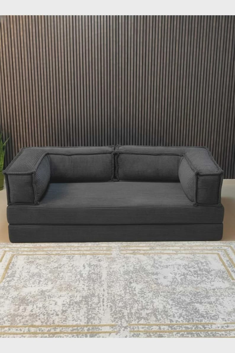 Peut inclure: Un canap&eacute; en velours c&ocirc;tel&eacute; gris fonc&eacute; avec deux accoudoirs et deux coussins de dossier. Le canap&eacute; repose sur une base pli&eacute;e. Le canap&eacute; est sur un tapis &agrave; motifs.