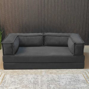 Peut inclure: Un canap&eacute; en velours c&ocirc;tel&eacute; gris fonc&eacute; avec deux accoudoirs et deux coussins de dossier. Le canap&eacute; repose sur une base pli&eacute;e. Le canap&eacute; est sur un tapis &agrave; motifs. L'arri&egrave;re-plan est un mur &agrave; panneaux de bois verticaux.