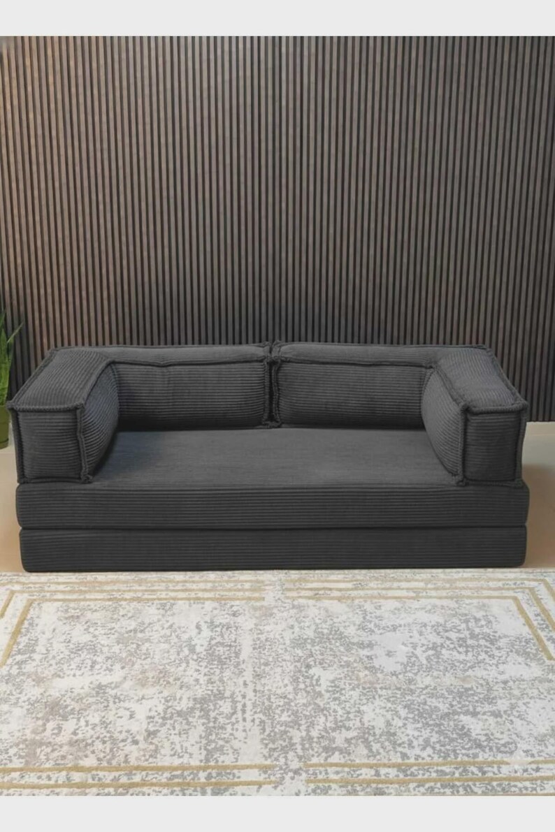 Peut inclure: Un canap&eacute; en velours c&ocirc;tel&eacute; gris fonc&eacute; avec deux accoudoirs et deux coussins de dossier. Le canap&eacute; repose sur une base pli&eacute;e. Le canap&eacute; est sur un tapis &agrave; motifs. L'arri&egrave;re-plan est un mur &agrave; panneaux de bois verticaux.