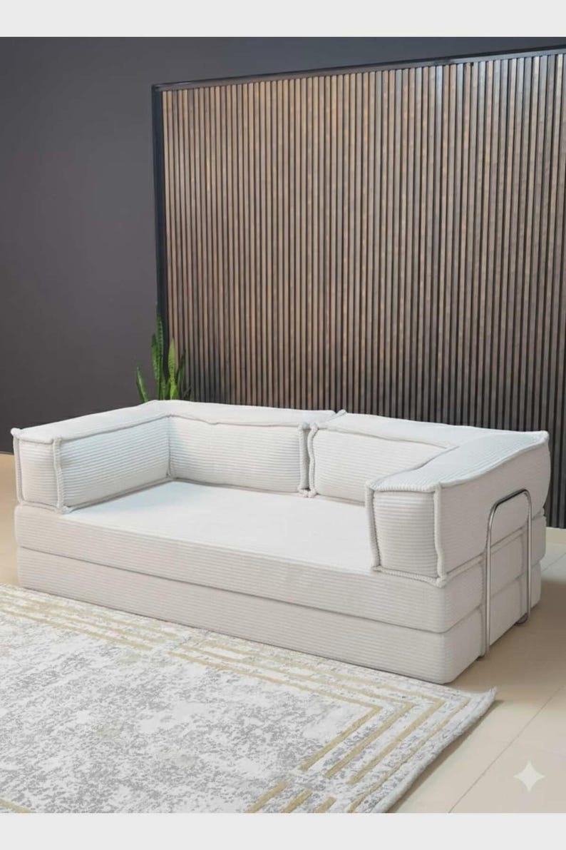 Pu&ograve; includere: Un moderno divano in velluto a coste bianco con un profilo basso. Il divano ha una forma rettangolare con braccioli e schienali spessi e imbottiti. Poggia su un tappeto chiaro con un motivo geometrico. Lo sfondo presenta una parete a doghe di legno.