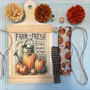 May include: A wooden cutting board with a printed design of pumpkins, apples, and a watering can. The design includes the text "Farm Fresh Pick Your Own! Cider Apples & Hay Rides". The board is surrounded by craft supplies, including brown twine, a black and white checkered ribbon, a burlap ribbon with a pumpkin print, and brown wooden beads.