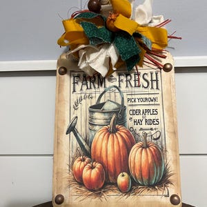 May include: Wooden sign with a fall theme. The sign features a vintage-style illustration of pumpkins, a watering can, and the text "Farm Fresh." The sign is adorned with a decorative bow in shades of yellow, green, and cream.