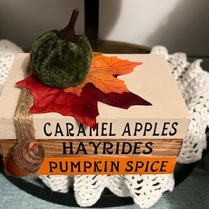 May include: A stack of three wooden blocks with fall-themed text. The top block is white with the text "CARAMEL APPLES". The middle block is brown with the text "HAYRIDES". The bottom block is orange with the text "PUMPKIN SPICE". A green velvet pumpkin and a red and orange fall leaf are on top of the blocks.