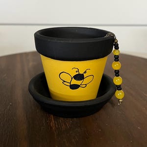 May include: A small yellow terracotta plant pot with a black rim and matching black saucer. The pot features a hand-painted black bee illustration on the front. A decorative chain of alternating black and yellow beads hangs from the rim.
