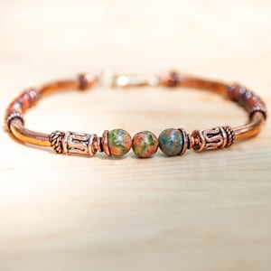 Pode incluir: Uma pulseira de cobre com três contas redondas e salpicadas em tons de verde e vermelho. A pulseira tem detalhes decorativos em cobre e um fecho prateado. A pulseira é exibida em uma superfície de madeira clara.