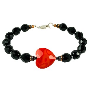 May include: A beaded bracelet featuring a central faceted red heart-shaped bead flanked by small metallic copper-toned spacers and dark gray beads. The rest of the bracelet consists of round, faceted black beads with a silver-toned metal clasp.
