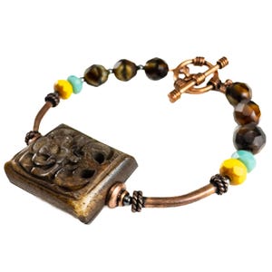 May include: A copper bracelet with a carved wooden charm and tiger's eye beads. The beads are brown, yellow, and turquoise.
