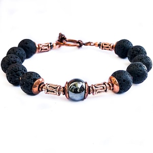 Könnte beinhalten: Ein Armband aus schwarzen Lavasteinen und Hämatitperlen mit kupfernen Akzenten. Das Armband hat einen Toggle-Verschluss.