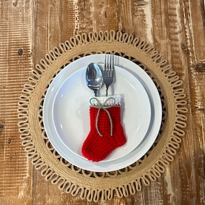 May include: A festive table setting featuring a white plate with a red crocheted stocking, spoon, and fork. The plate rests on a woven placemat with a decorative edge, all set on a wooden table. Christmas-themed decor.