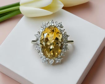 May include: A silver ring with a large, oval-cut yellow gemstone, surrounded by smaller, clear stones. The ring is set on a white surface, with two white tulips in the background. The ring is a piece of jewelry.