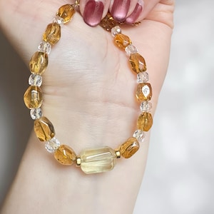 May include: A beaded bracelet featuring a mix of amber and clear crystal beads. The bracelet has a central rectangular amber stone and gold-colored metal accents. The beads are strung together, creating a delicate and elegant piece of jewelry.