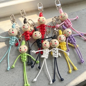 Puede incluir: Un conjunto de nueve llaveros de macramé, cada uno con una cabeza de cuenta de madera redonda con una cara sonriente pintada y mejillas rosadas. Los cuerpos están hechos de cordón anudado de colores azul claro, rojo, negro, blanco, verde, amarillo y morado, con cierres de mosquetón de metal plateado unidos a la parte superior de cada figura.