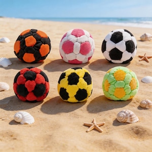 May include: Six colorful, plush soccer balls in various color combinations, including black and orange, pink and white, and black and white, are arranged on a sandy beach. Other colors include red and black, yellow and black, and green and yellow. Seashells and starfish are scattered around.
