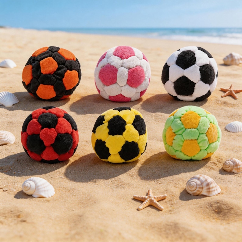 May include: Six colorful, plush soccer balls in various color combinations, including black and orange, pink and white, and black and white, are arranged on a sandy beach. Other colors include red and black, yellow and black, and green and yellow. Seashells and starfish are scattered around.
