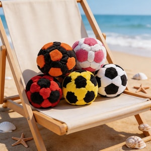 May include: Five colorful soccer balls in various colors, including black and orange, pink and white, red and black, yellow and black, and black and white. The balls are on a beach chair with a beach and ocean background.