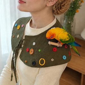 May include: An olive green fabric shoulder cape adorned with various colorful buttons in shades of yellow, red, blue, and orange. A small yellow, orange, and green parrot is perched on the cape, holding a small wooden dowel in its beak.