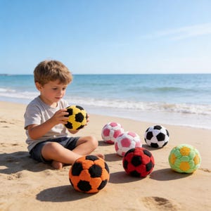 May include: A young child sits on a sandy beach, surrounded by colorful soccer balls. The balls are in various colors, including yellow, black, orange, red, pink, white, and green. The ocean and blue sky are in the background.