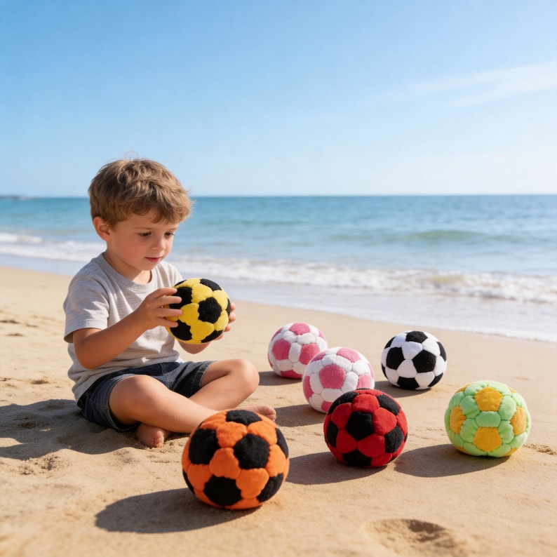 May include: A young child sits on a sandy beach, surrounded by colorful soccer balls. The balls are in various colors, including yellow, black, orange, red, pink, white, and green. The ocean and blue sky are in the background.