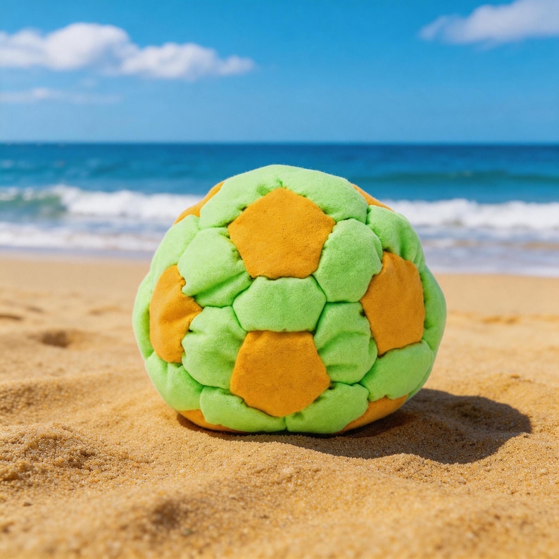 May include: A colorful, soft soccer ball made of green and orange fabric, resting on a sandy beach. The ball's design features a pattern of pentagons and hexagons. The ocean and blue sky are visible in the background.