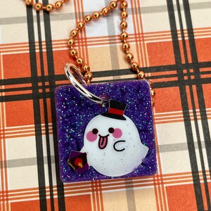 May include: A Halloween-themed pendant necklace. The square pendant features a cute ghost with a top hat and a lantern against a purple glitter background. The necklace has a copper-colored ball chain.