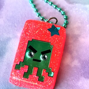 May include: A pink glitter resin pendant necklace with a green pixelated alien face and a silver star. The necklace has a silver chain.