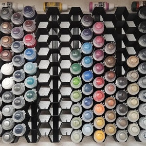 May include: A black hexagonal paint storage rack with rows of small paint bottles in various colours. The bottles are arranged in a grid pattern, with each row containing a different colour.