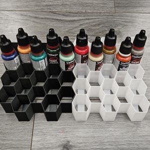 May include: A black and white honeycomb paint bottle organiser with 12 bottles of paint in various colours. The bottles are labelled with the brand "Vallejo" and "Game Color".