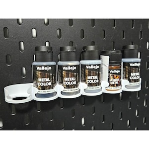 May include: A selection of Vallejo Metal Colour acrylic paints in metallic grey and bronze shades, held in white supports. The bottles are labelled with the brand and product information. The paints are mounted on a black pegboard.