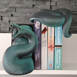 May include: A pair of green snake bookends holding up a stack of books. The books are titled "Nine Perfect Strangers", "The Devil Wears Prada", "Paper Palace", and "The Seers".