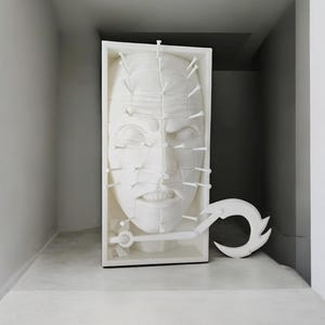 May include: A white sculpture of a face with nails protruding from the surface, displayed in a white frame. A white hook is attached to the bottom of the frame.