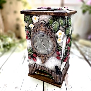 May include: A decorative clock with a rectangular shape, featuring a vintage design. The clock face displays Roman numerals and is surrounded by a floral motif of strawberries, white flowers, and green leaves. The clock is painted in a distressed style.