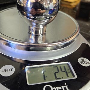 May include: A silver metal sphere with a reflective surface is placed on a digital kitchen scale. The scale reads 1.24 ounces. The scale is black with a silver trim and the brand name "Ozeri" is printed on the front.