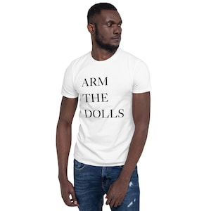 May include: White t-shirt with the text "ARM THE DOLLS" in black, displayed in a stacked format. The shirt is worn with blue jeans. The image shows a person wearing the shirt.
