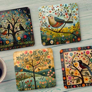 May include: Four square coasters with colorful bird and tree illustrations. One coaster features a tree with multiple birds, another shows a bird on a branch, and two others depict trees with birds and flowers. A red mug of coffee is in the corner.
