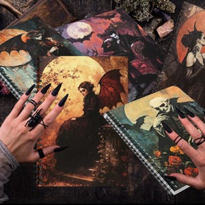 Könnte beinhalten: Vier spiralgebundene Notizbücher mit gotisch inspirierten Illustrationen. Die Cover zeigen eine Fledermaus, eine Frau mit Flügeln, ein Paar und ein Skelett. Die Notizbücher liegen auf einer dunklen Oberfläche mit einer Hand, die schwarzen Nagellack und Ringe trägt.
