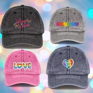 Könnte beinhalten: Vier Baseballkappen mit gestickten Designs. Die Kappen sind in verschiedenen Farben erhältlich: Schwarz, Grau, Rosa und Marineblau. Die Designs umfassen den Text "Love is Love" und "LOVE" mit Regenbogenfarben und Regenbogenherzen.