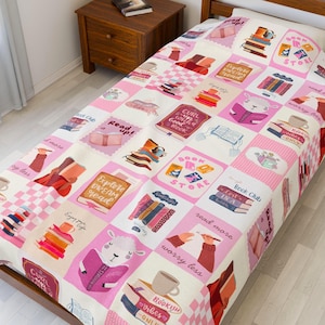 Könnte beinhalten: Eine rosa-weiß gemusterte Bettdecke mit Illustrationen zum Thema Bücher. Das Design umfasst Bilder von Bücherstapeln, Kaffeetassen und einem lesenden Schaf. Der Text enthält "Read up!", "Book Store" und "Explore Dream Read".