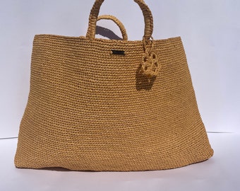 May include: A large, tan woven tote bag with two woven handles. The bag has a small woven charm attached to one of the handles.