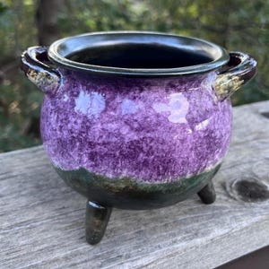 May include: A small, round ceramic cauldron with three short legs and two side handles. The pot features a glossy finish with a vibrant purple upper section that transitions into a dark, mottled green and black base, with a dark interior rim.