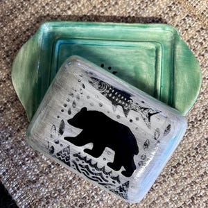May include: A ceramic butter dish consisting of a rectangular green base with handles and a matching lid. The lid features a white brushed finish with a black silhouette of a bear, a fish skeleton, and geometric patterns printed on the surface.