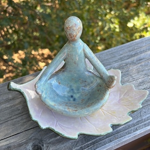 May include: A ceramic sculpture of a figure in a seated meditation pose, featuring a glossy light blue glaze with brown speckles. The figure is attached to a base shaped like a large, flat leaf with a pale pink and cream iridescent finish.