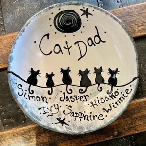 May include: A hand-painted ceramic plate with a whimsical design. The plate features a black cat silhouette design with the words "Cat Dad" and the names of the cats: Simon, Ivy, Jasper, Sapphire, Hisako, and Winnie. The plate has a white and blue color scheme.