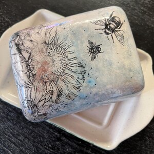 May include: A rectangular ceramic butter dish with a lid featuring a black line art illustration of a large sunflower and two bees. The lid has a light blue and white mottled glaze finish. The base is a plain, off-white ceramic tray with edges.